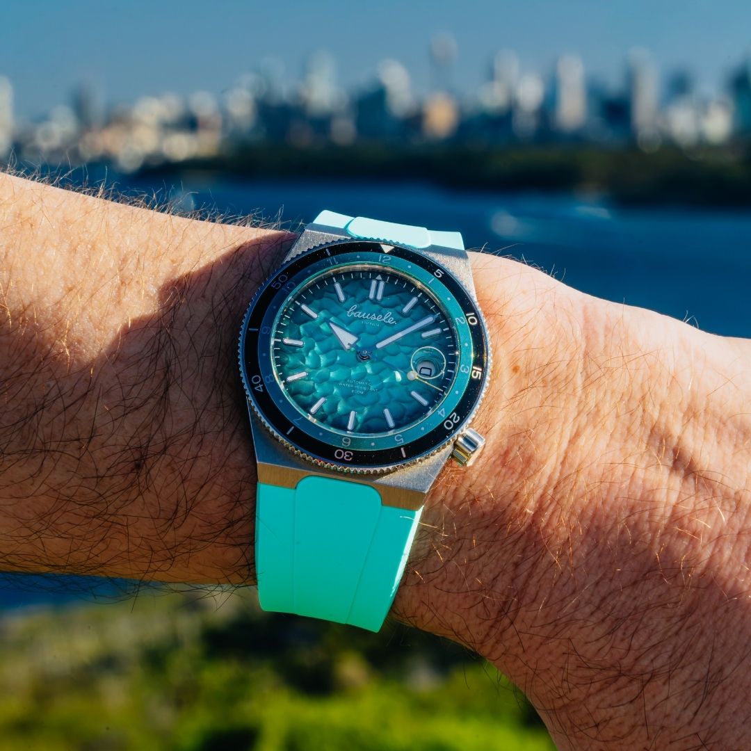 A close-up of a wrist wearing a turquoise and silver watch with a matching turquoise strap, set against a blurred outdoor background of water and a city skyline.